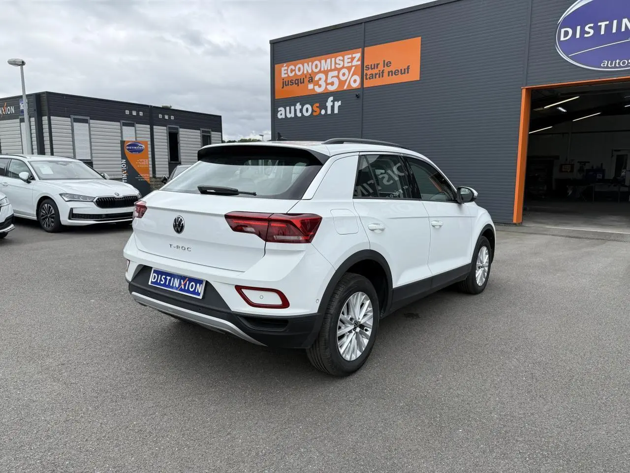 Vue 3/4 arrière droite d'un Volkswagen T-Roc blanc pur garé devant un concessionnaire avec panneaux publicitaires visibles.