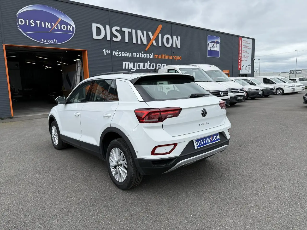 Volkswagen T-Roc blanc pur vu en 3/4 arrière droit, garé devant un bâtiment avec plusieurs véhicules blancs alignés.