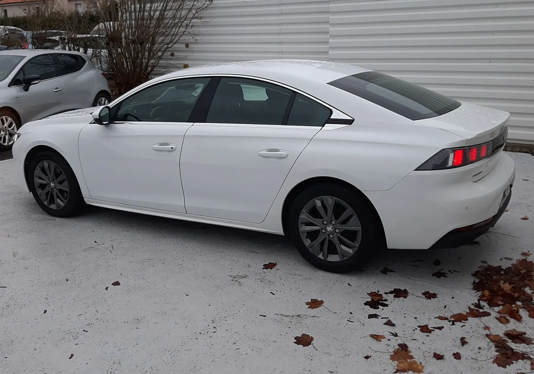 Peugeot 508 blanc vue de profil côté gauche, avec feux arrière allumés et jantes foncées, stationnée sur sol clair.