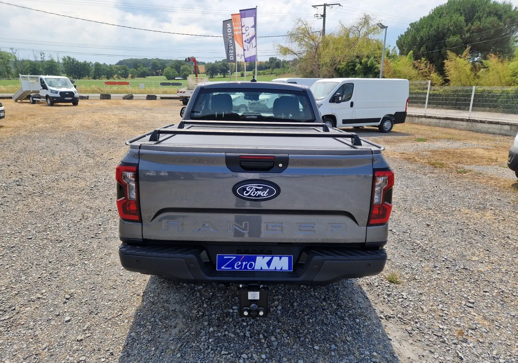 Vue arrière d'un Ford Ranger Super Cabine gris carbone avec rideau de plateau rigide et attelage visible sur un terrain gravillonné.