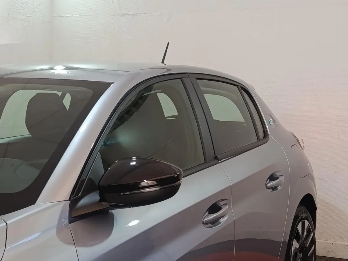 Vue côté gauche avant d'une Peugeot 208 électrique gris clair avec rétroviseur noir brillant et poignées de porte assorties.