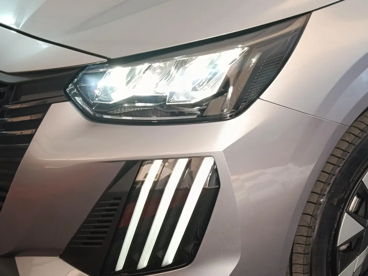 Gros plan sur l'avant droit gris clair de la Peugeot 208 électrique 2024, mettant en valeur les phares LED et les feux diurnes verticaux.