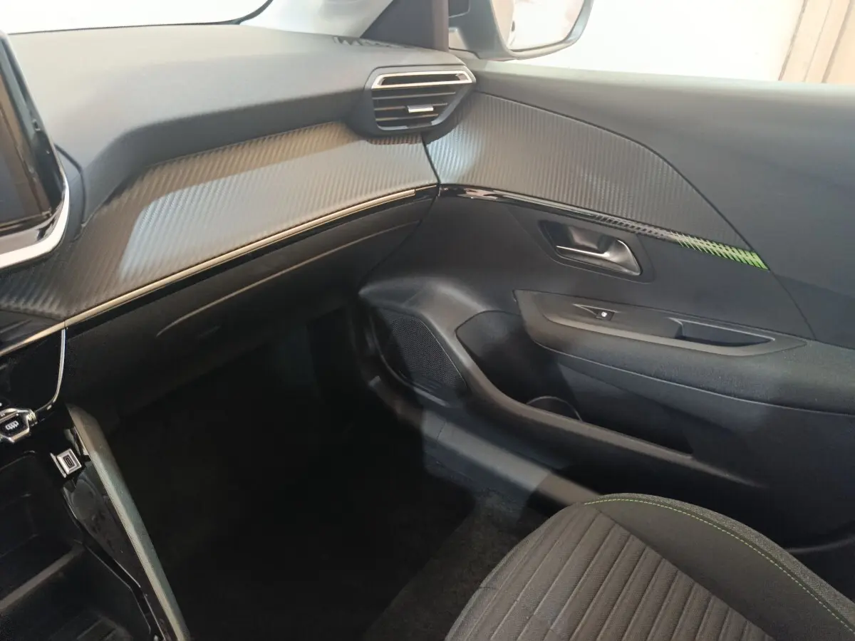 Intérieur côté passager du Peugeot 208 électrique 2024, tableau de bord noir avec insert effet carbone et surpiqûres vertes sur siège.