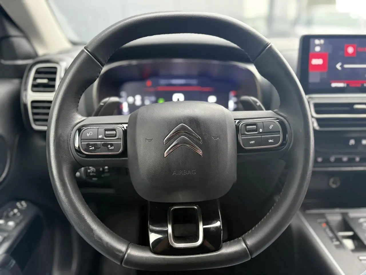 Vue rapprochée du volant en cuir noir du Citroën C5 Aircross 2024 avec commandes intégrées et tableau de bord numérique flou.