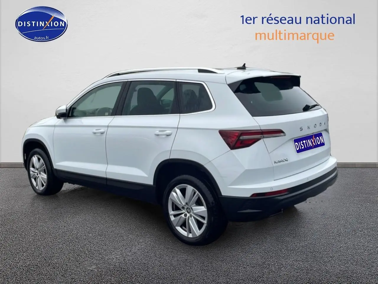 Vue 3/4 arrière droite d'un Skoda Karoq blanc glacial avec jantes alu et barres de toit visibles.