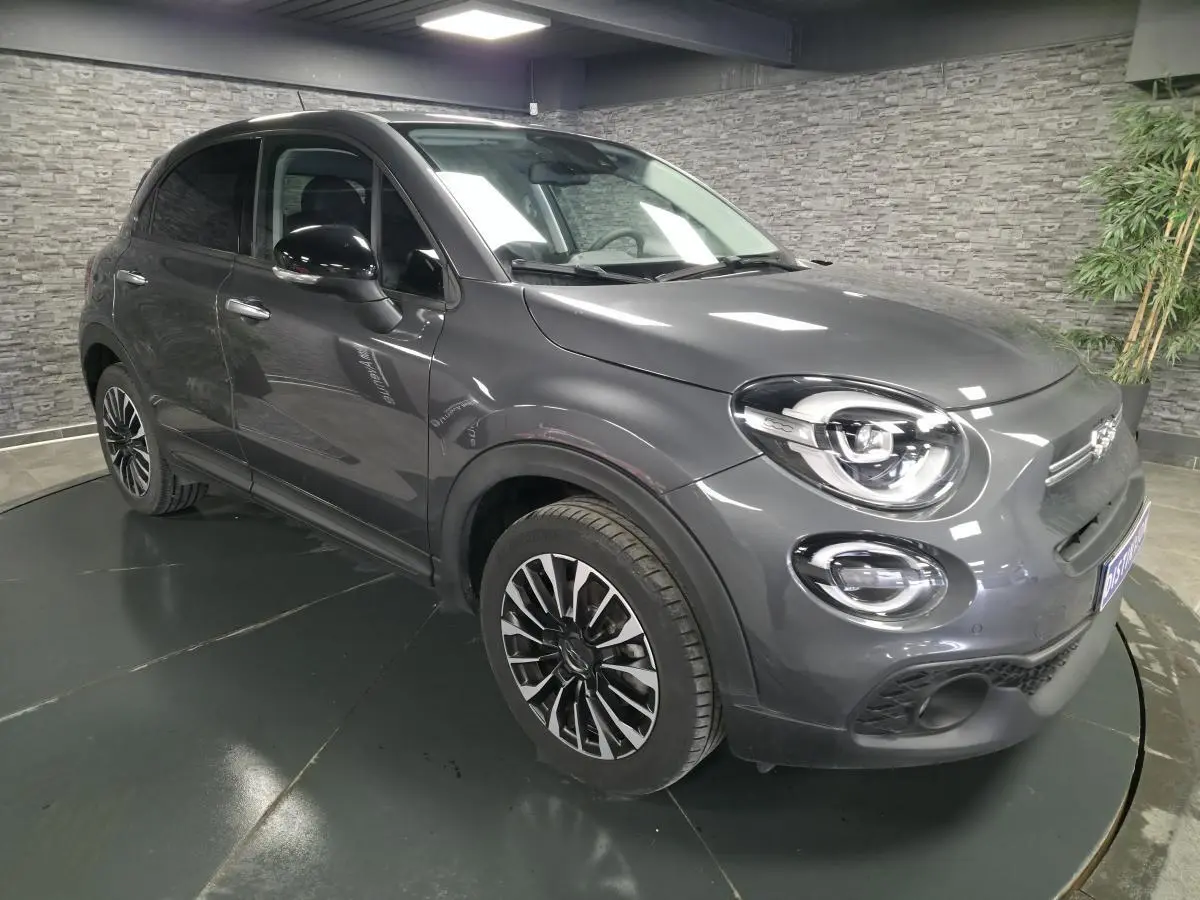 Fiat 500X gris en 3/4 avant droit, avec jantes alliage 17'' et feux LED distinctifs dans un showroom.