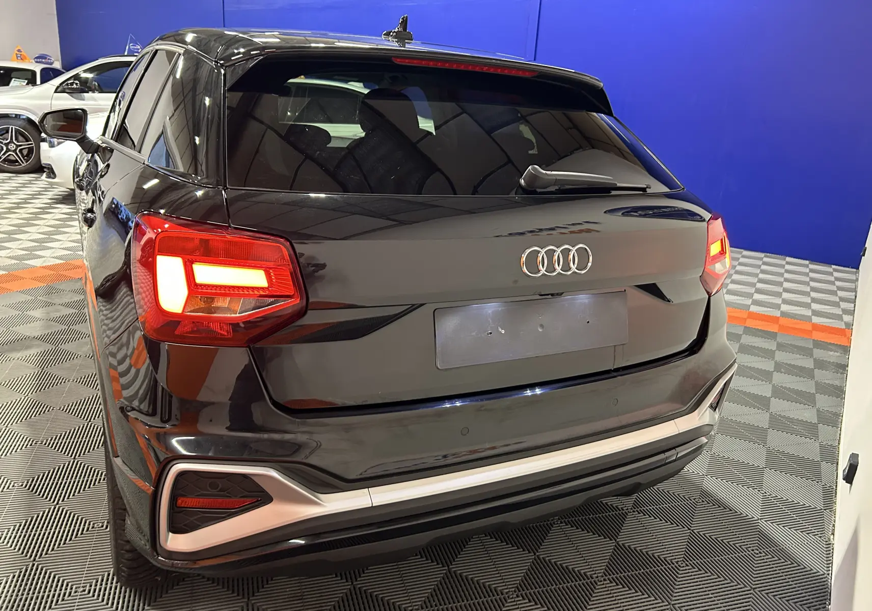 Vue 3/4 arrière d'un Audi Q2 noir métal 2023 avec feux arrière allumés et hayon électrique en intérieur showroom.