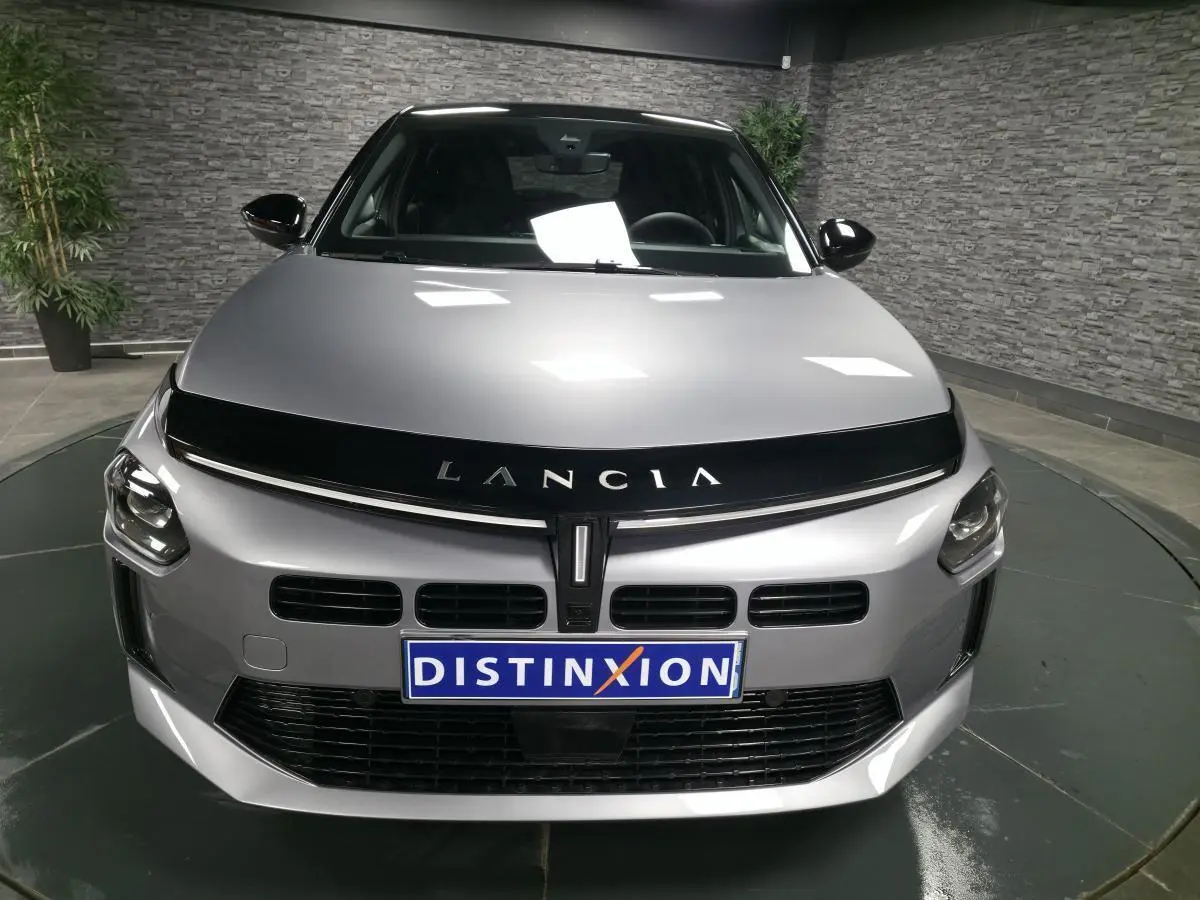 Vue avant d'une Lancia Ypsilon 2025 gris métallisé avec calandre noire et toit bi-color noir en intérieur showroom.