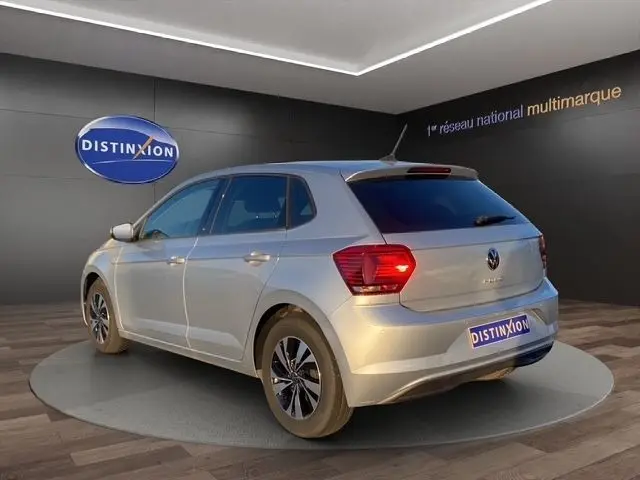 Volkswagen Polo gris clair vue 3/4 arrière droit en intérieur avec feux arrière allumés et jantes noires.
