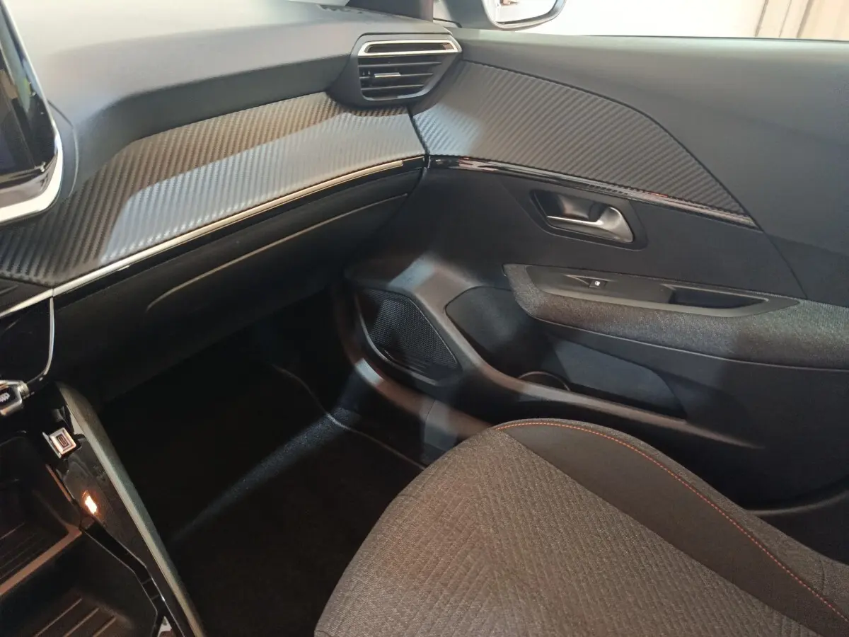 Vue intérieure côté passager de la Peugeot 208 Hybrid 2025, tableau de bord et porte avec finition carbone gris clair.
