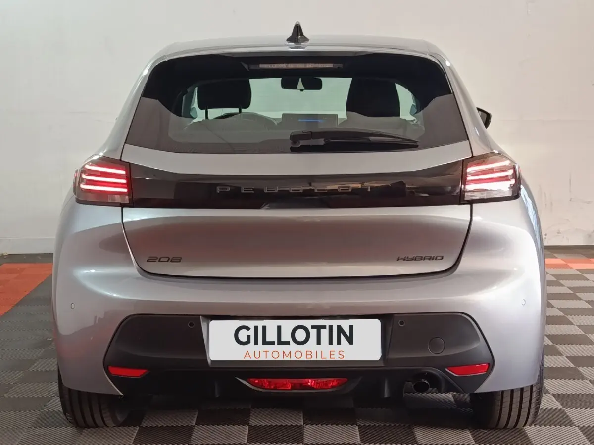 Vue arrière d'une Peugeot 208 Hybrid gris clair avec feux LED allumés sur fond intérieur neutre.