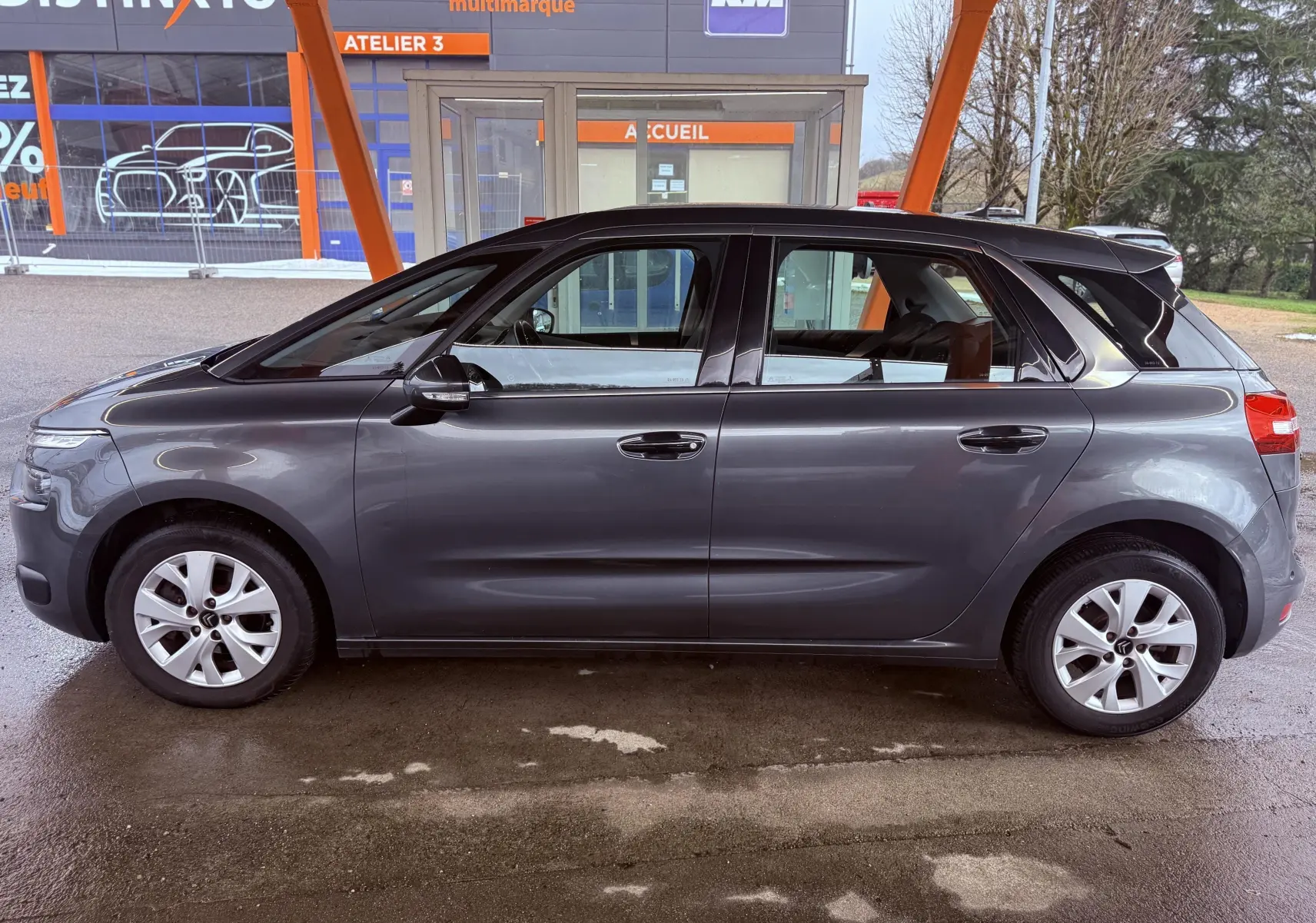 Vue de profil côté gauche d'une Citroën C4 Picasso gris platinium métallisé stationnée sous abri.