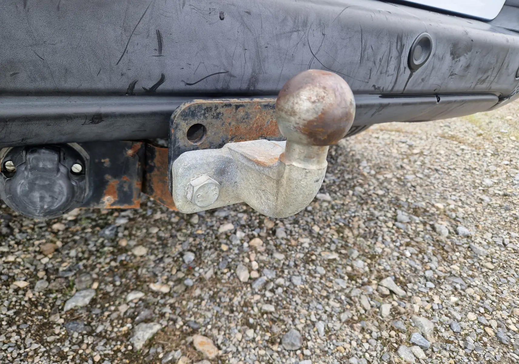 Gros plan sur l'attelage rouillé fixé au pare-chocs arrière noir du Peugeot Partner blanc, avec prise électrique visible.