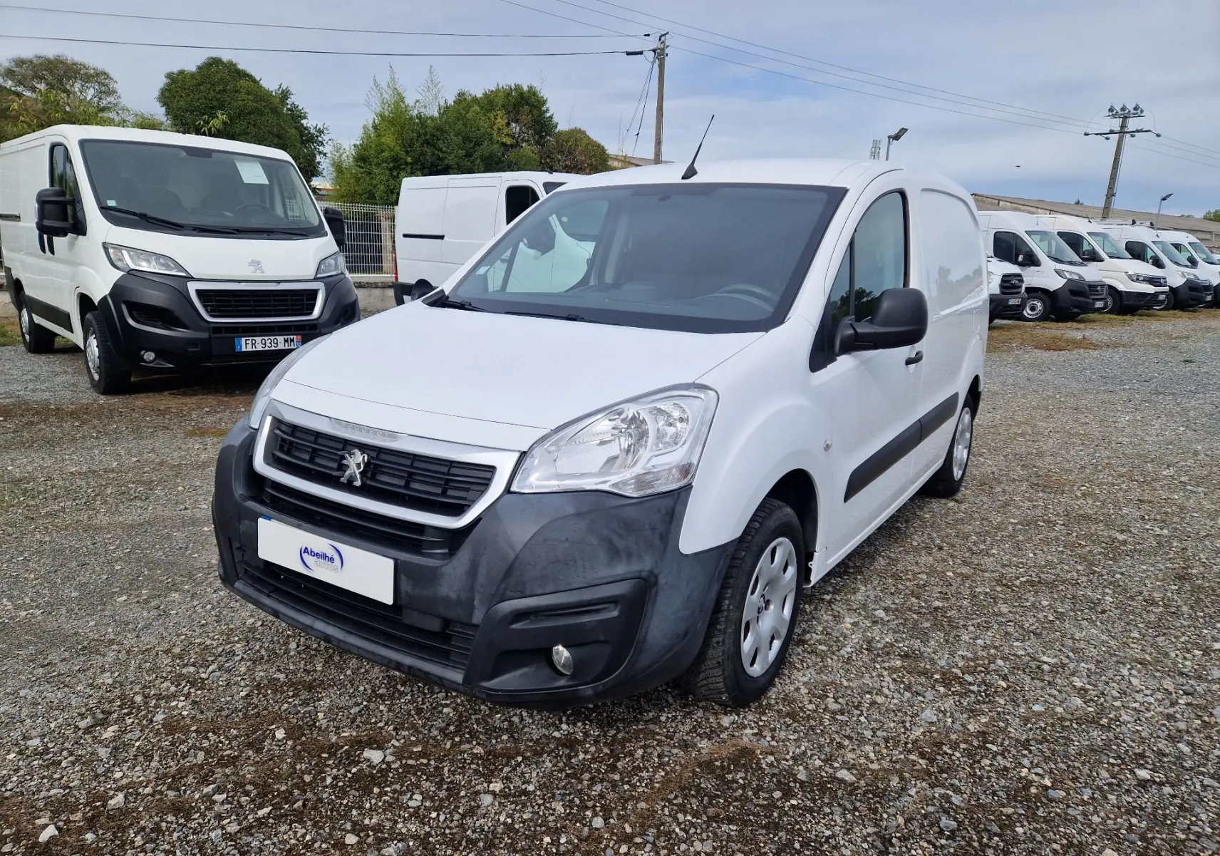 Vue 3/4 avant droit d’un Peugeot Partner blanc 2017 avec pare-chocs noir mat et calandre Peugeot visible sur un parking.