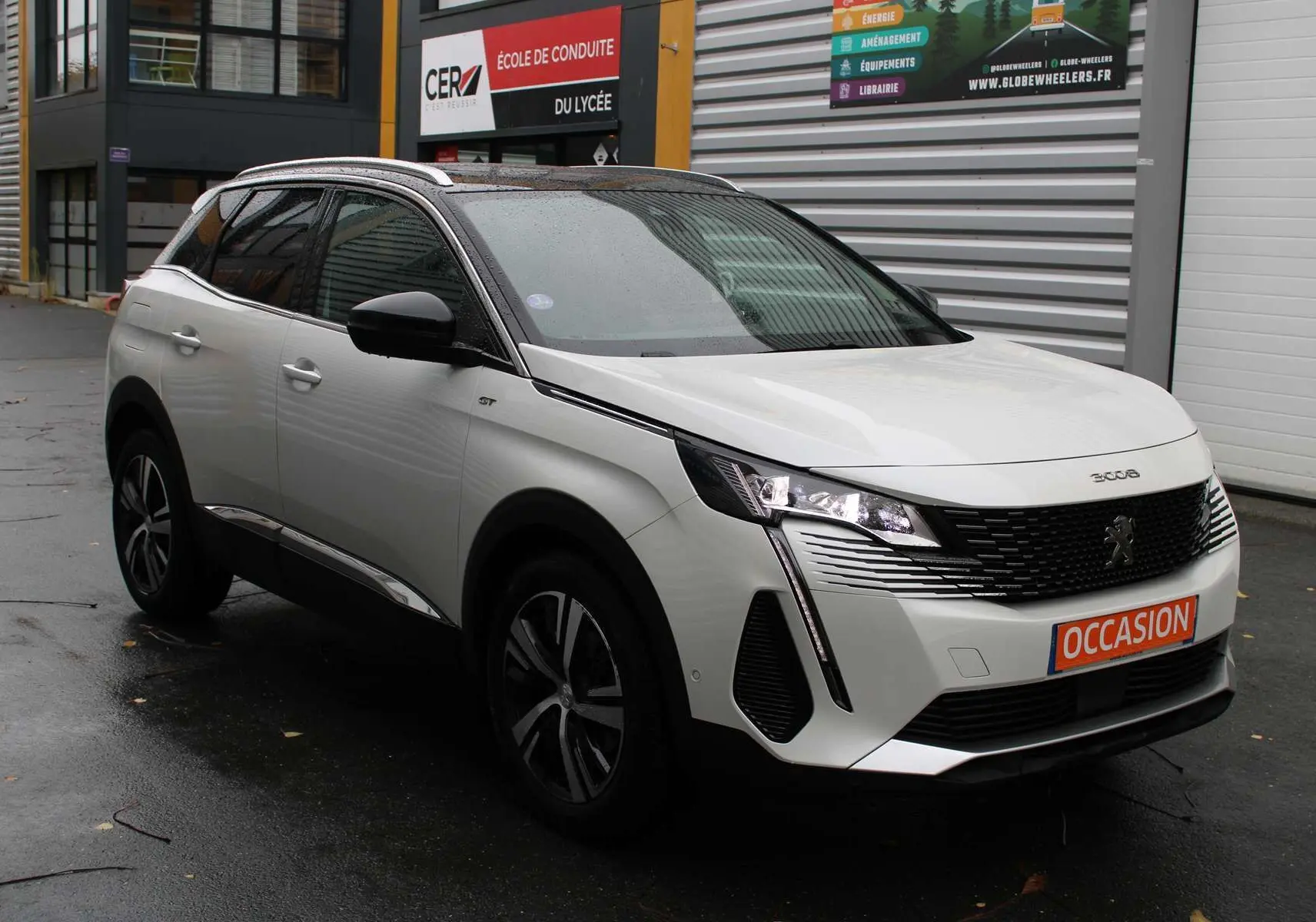 Peugeot 3008 blanc nacré en 3/4 avant droit avec toit panoramique ouvrant sous la pluie devant un garage.