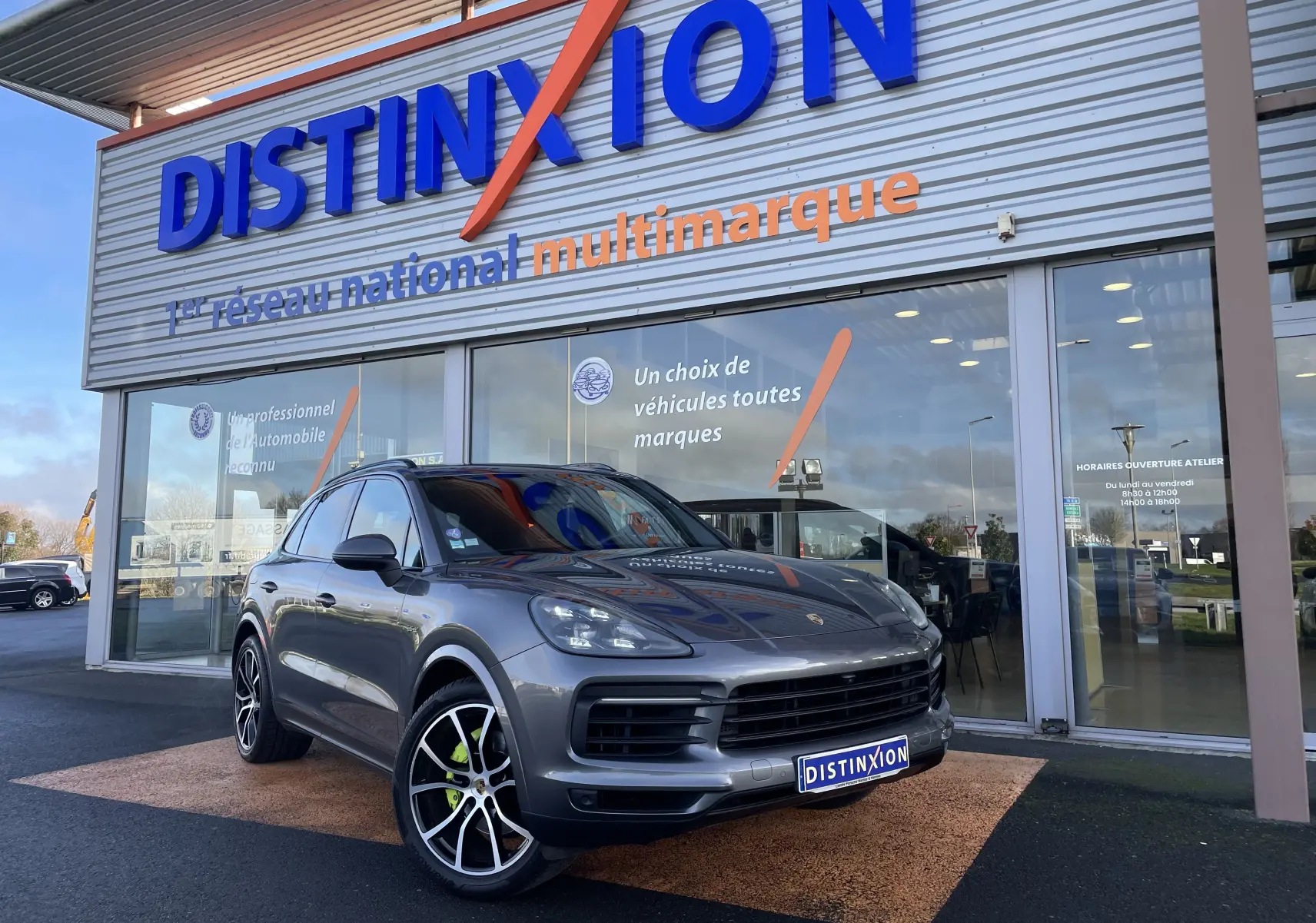 Porsche Cayenne E-Hybrid gris Kerguelen en 3/4 avant droit, roues avec étriers verts, devant un showroom Distinxion.