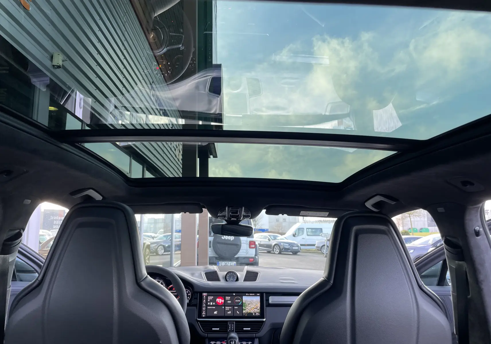 Vue intérieure avant du Porsche Cayenne E-Hybrid gris Kerguelen 2018 avec toit panoramique et sièges sport noirs.