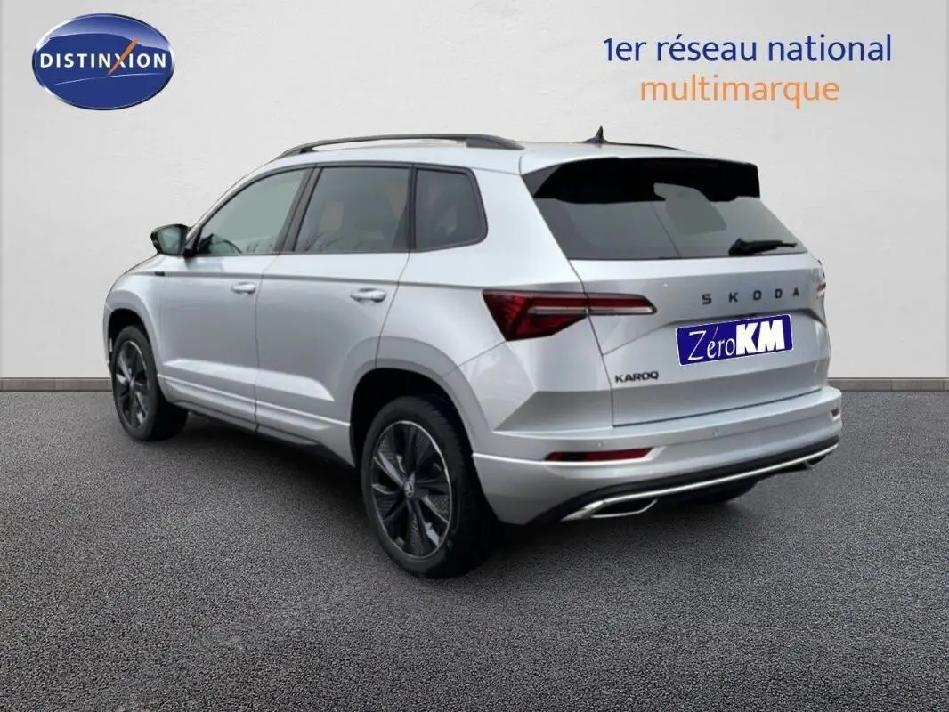 Vue 3/4 arrière gauche d'un Skoda Karoq argent avec barres de toit et jantes noires, sur fond neutre.