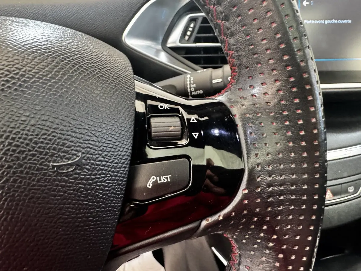 Gros plan sur le volant cuir noir perforé avec surpiqûres rouges du Peugeot 308 GT Line, commandes intégrées visibles.
