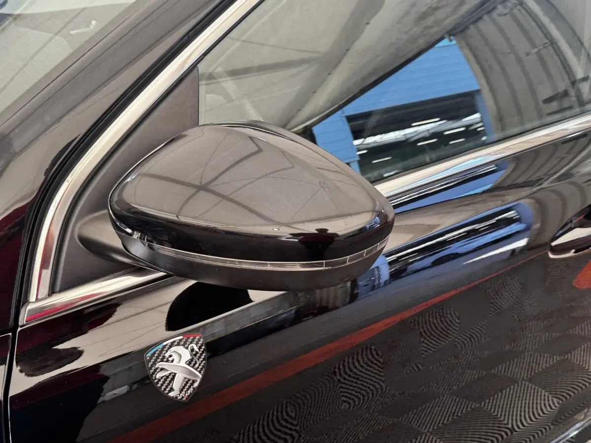 Gros plan sur le rétroviseur côté gauche noir brillant et l'écusson Peugeot en carbone sur la porte noire du Peugeot 308.