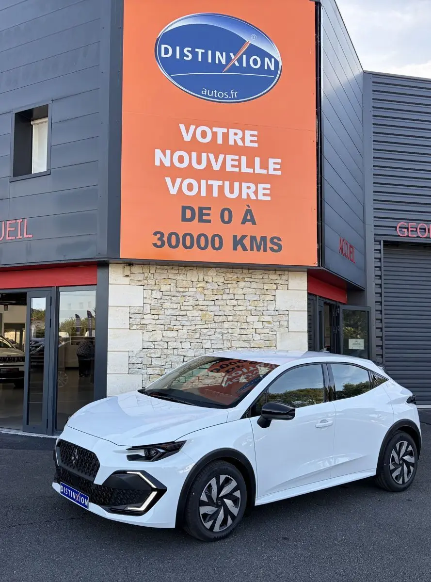 Renault Clio blanche vue en 3/4 avant droit, avec calandre noire et jantes au design moderne, devant un bâtiment commercial.