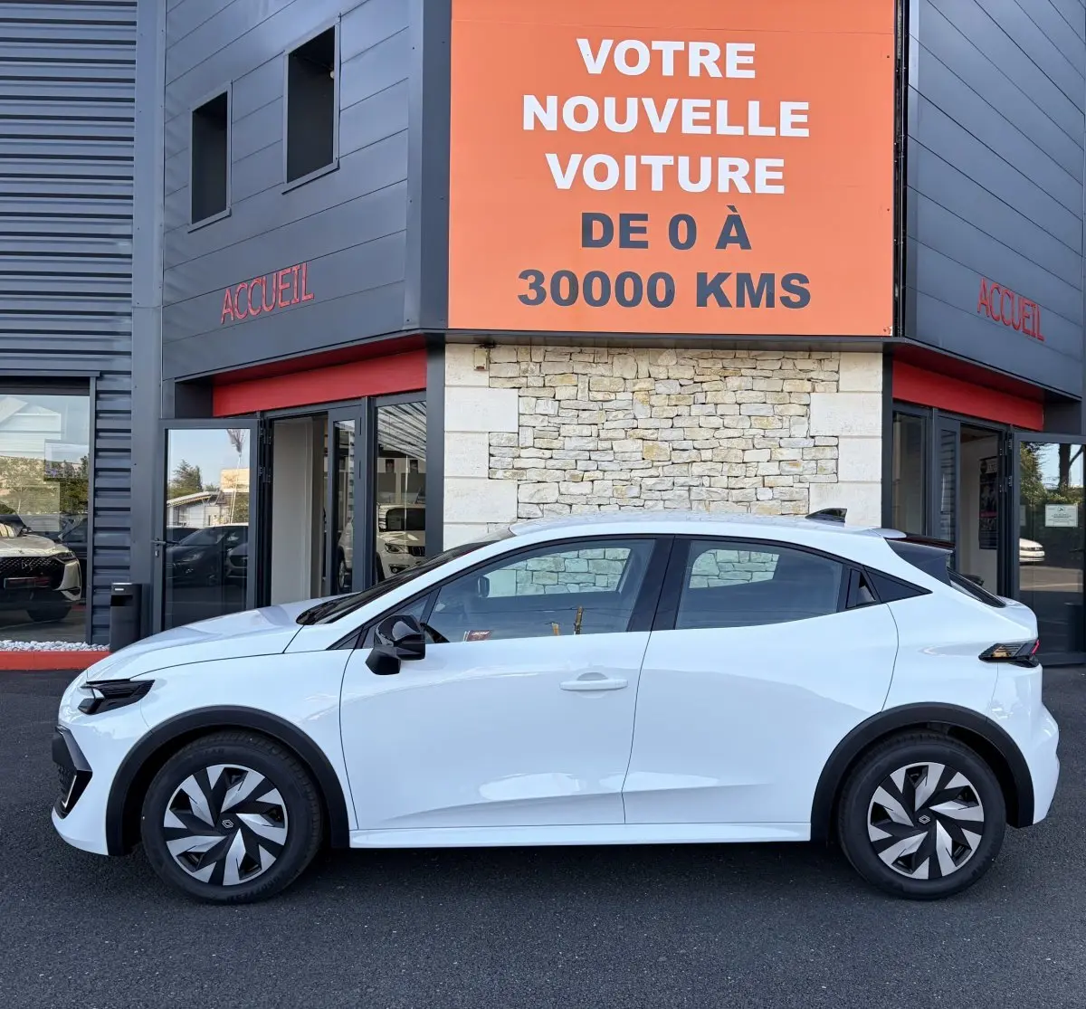 Profil côté gauche d'une Renault Clio blanche 2026 garée devant un bâtiment avec un panneau orange indiquant "votre nouvelle voiture de 0 à 30000 kms".