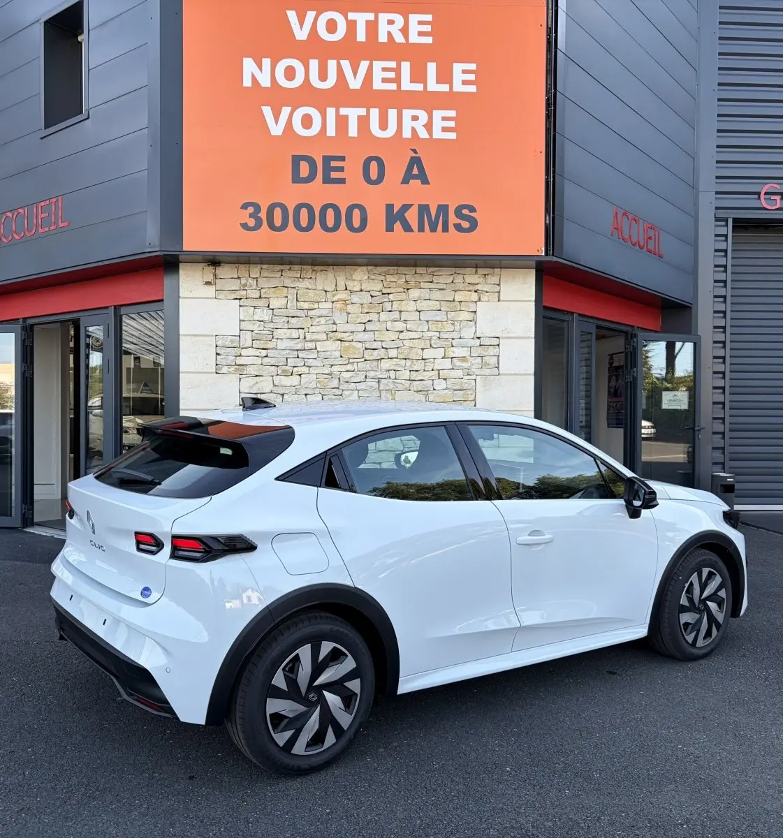 Renault Clio blanche vue 3/4 arrière droit, avec feux arrière LED et jantes au design moderne, devant un bâtiment commercial.
