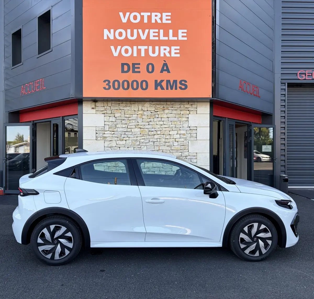 Profil droit d'une Renault Clio blanche 2026 garée devant un bâtiment avec une enseigne orange "Votre nouvelle voiture de 0 à 30000 kms".