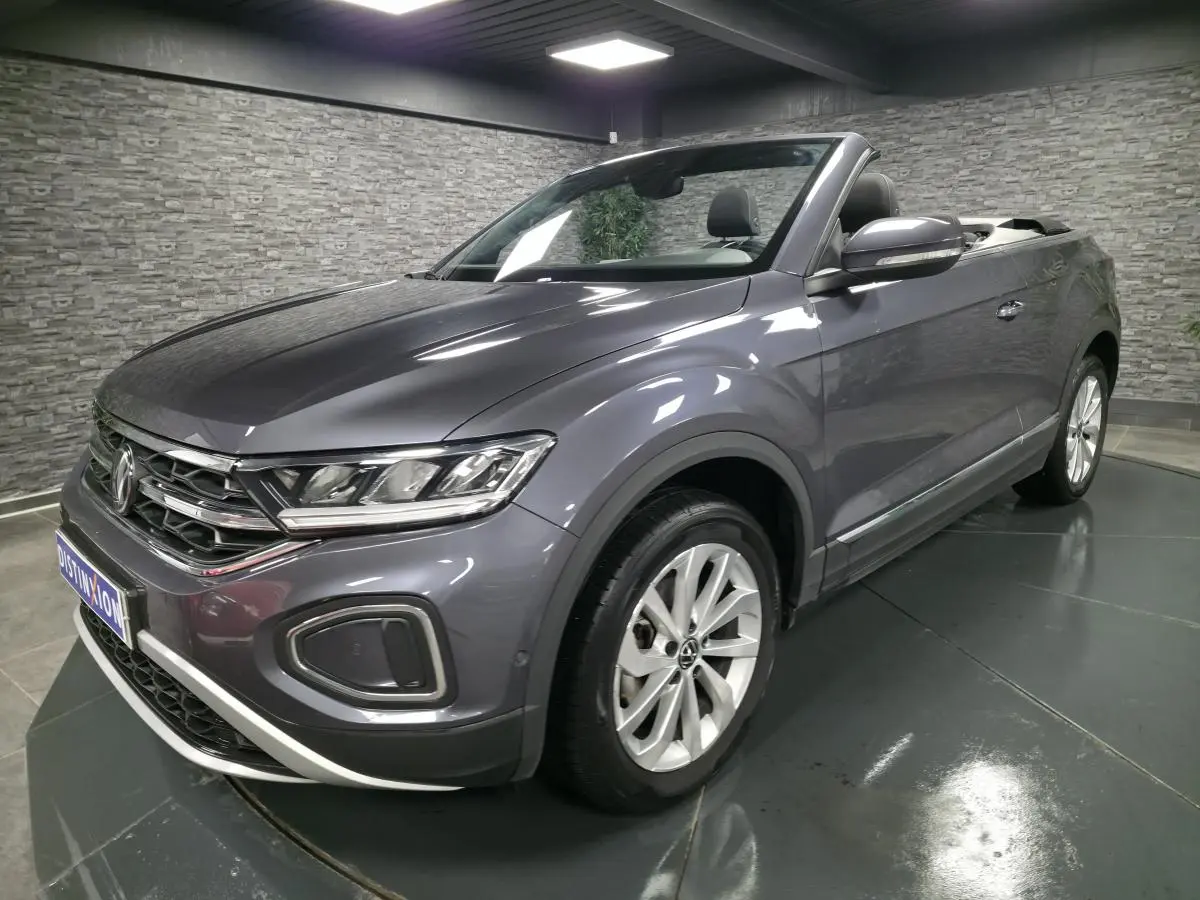 Volkswagen T-Roc cabriolet gris métallisé en 3/4 avant droit, capote ouverte et jantes alliage visibles.