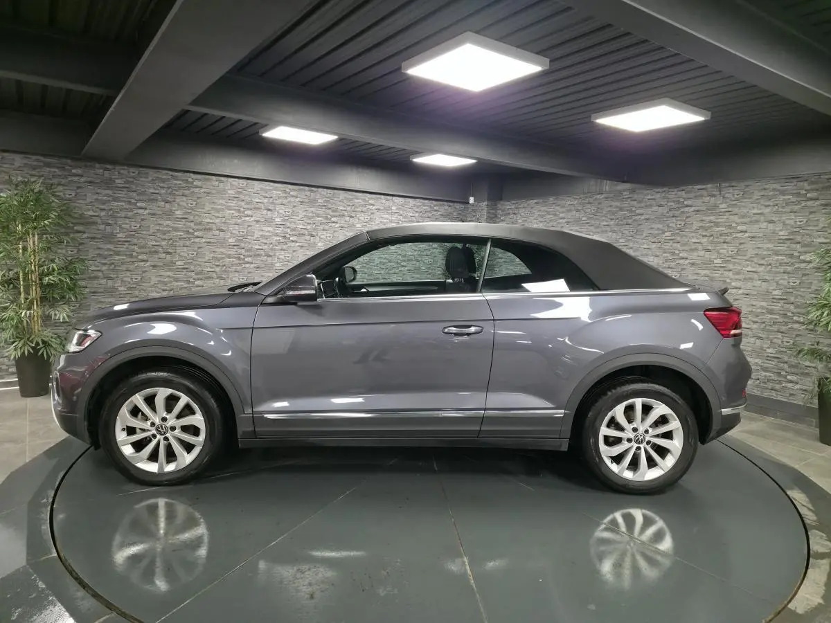Volkswagen T-Roc Cabriolet gris métallisé vue de profil côté gauche avec capote noire fermée en intérieur showroom.
