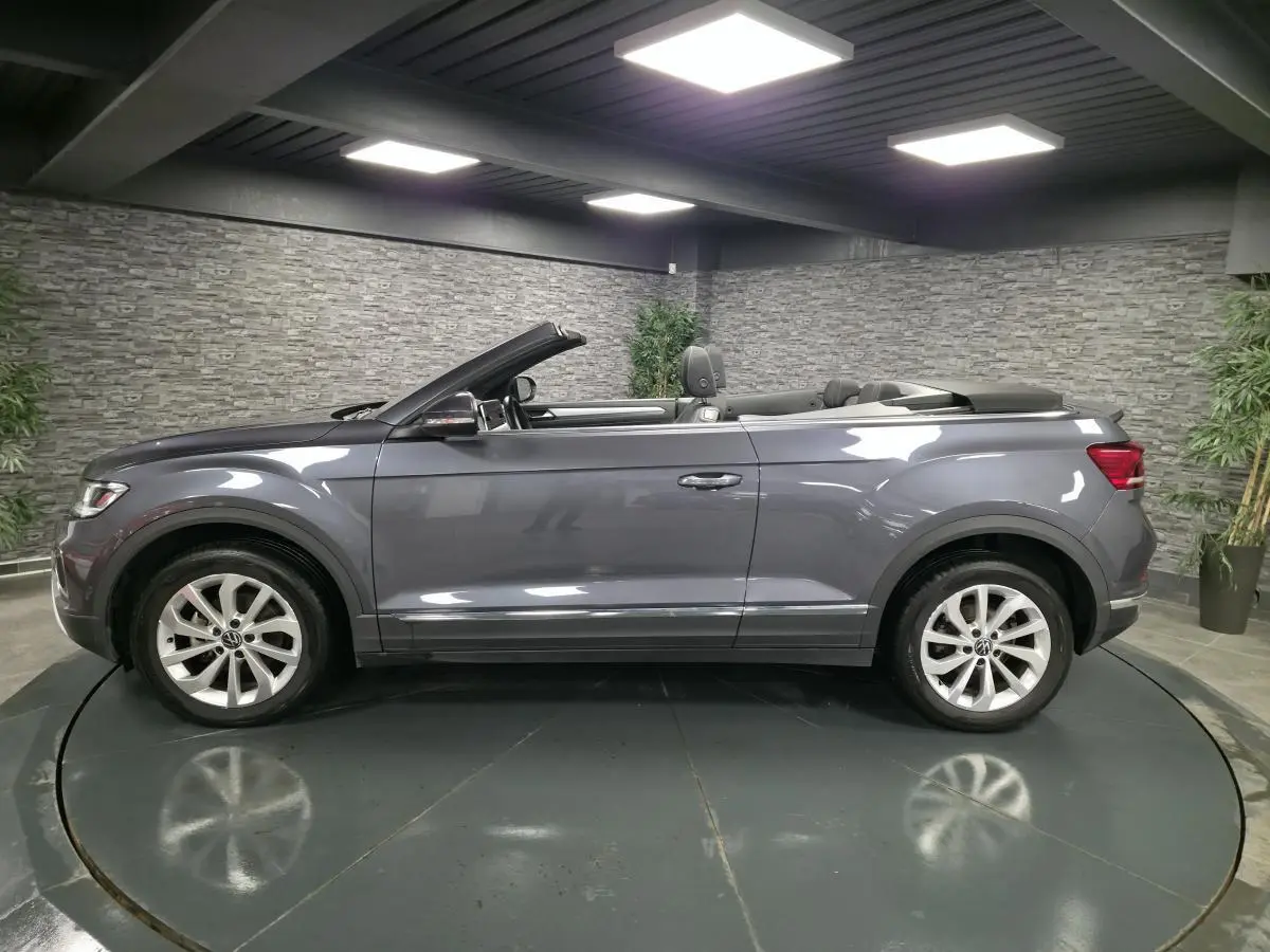 Vue de profil côté gauche d'un Volkswagen T-Roc Cabriolet gris métallisé avec capote ouverte en intérieur showroom.