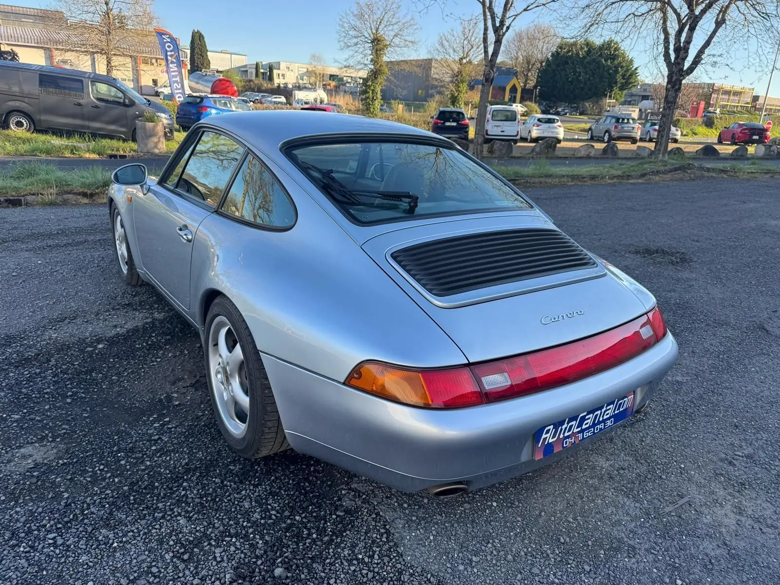 Vue 3/4 arrière droite d'une Porsche 911 Carrera 993 gris polaire avec jantes 17 pouces sur parking extérieur.