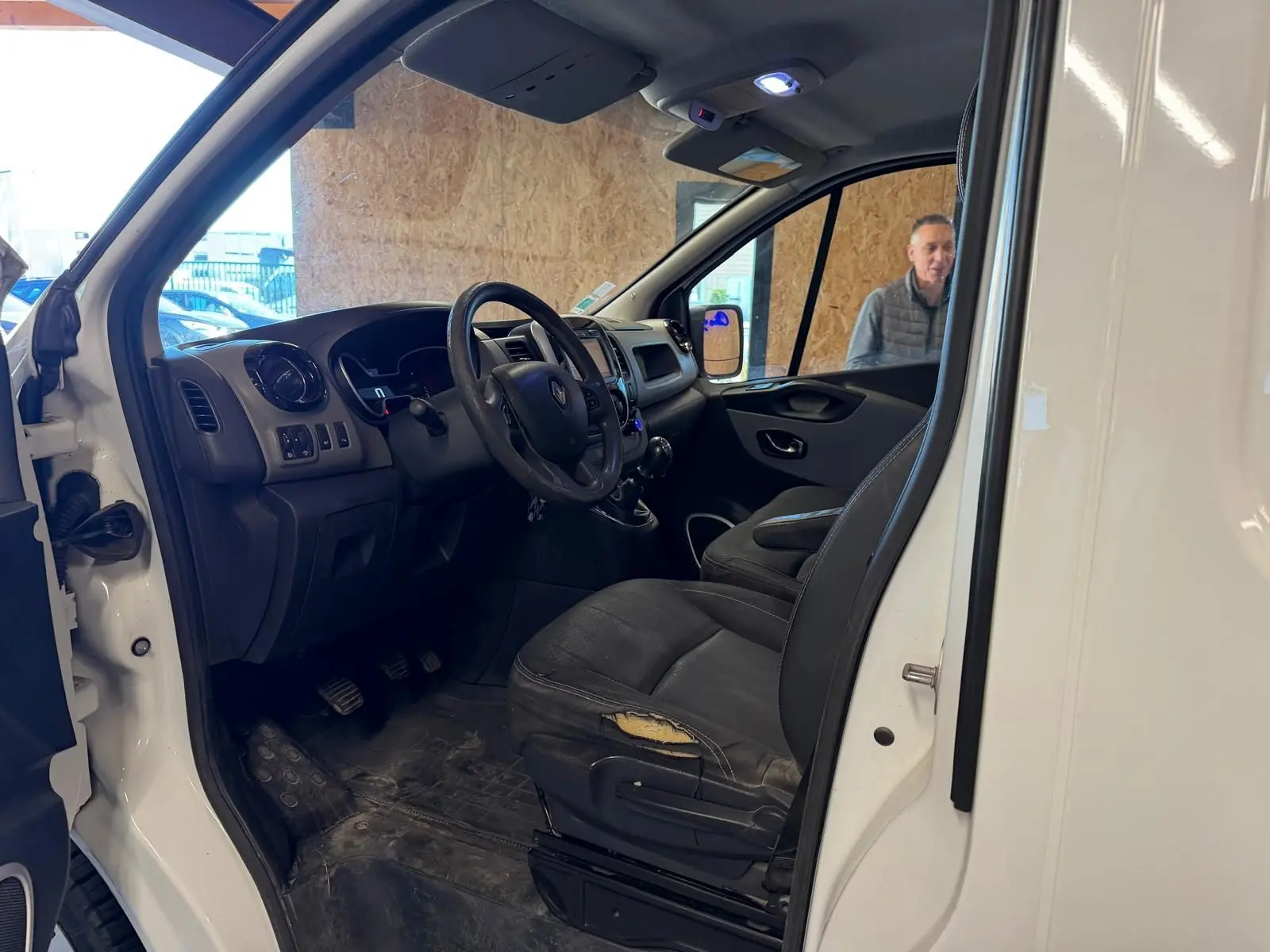 Vue intérieure côté conducteur du Renault Trafic blanc, montrant le tableau de bord et siège usé avec volant et levier de vitesse.