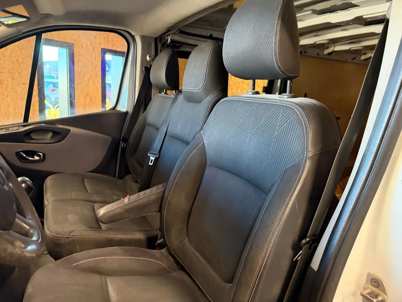 Vue intérieure côté gauche du Renault Trafic Fourgon 2017, sièges avant en tissu noir avec habillage bois visible en arrière-plan.