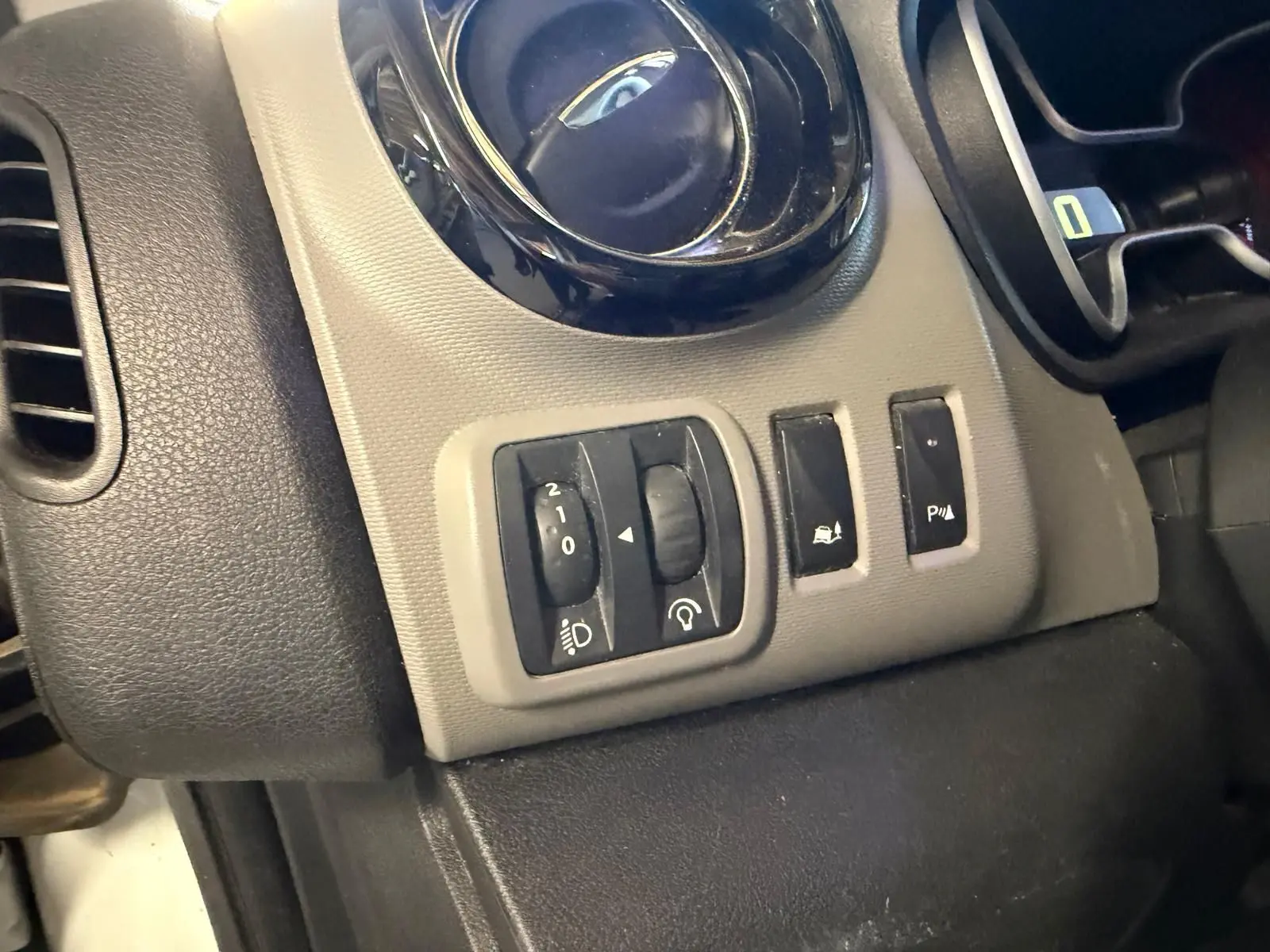 Gros plan sur les commandes de réglage des phares et aides à la conduite du tableau de bord gris et noir du Renault Trafic blanc.