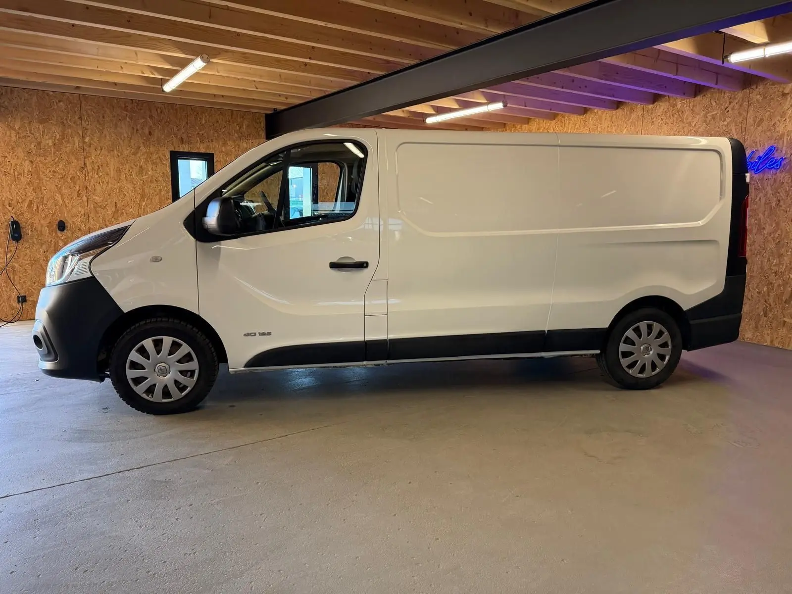 Vue de profil côté gauche d'un Renault Trafic Fourgon blanc 2017 avec habillage bois en arrière-plan.