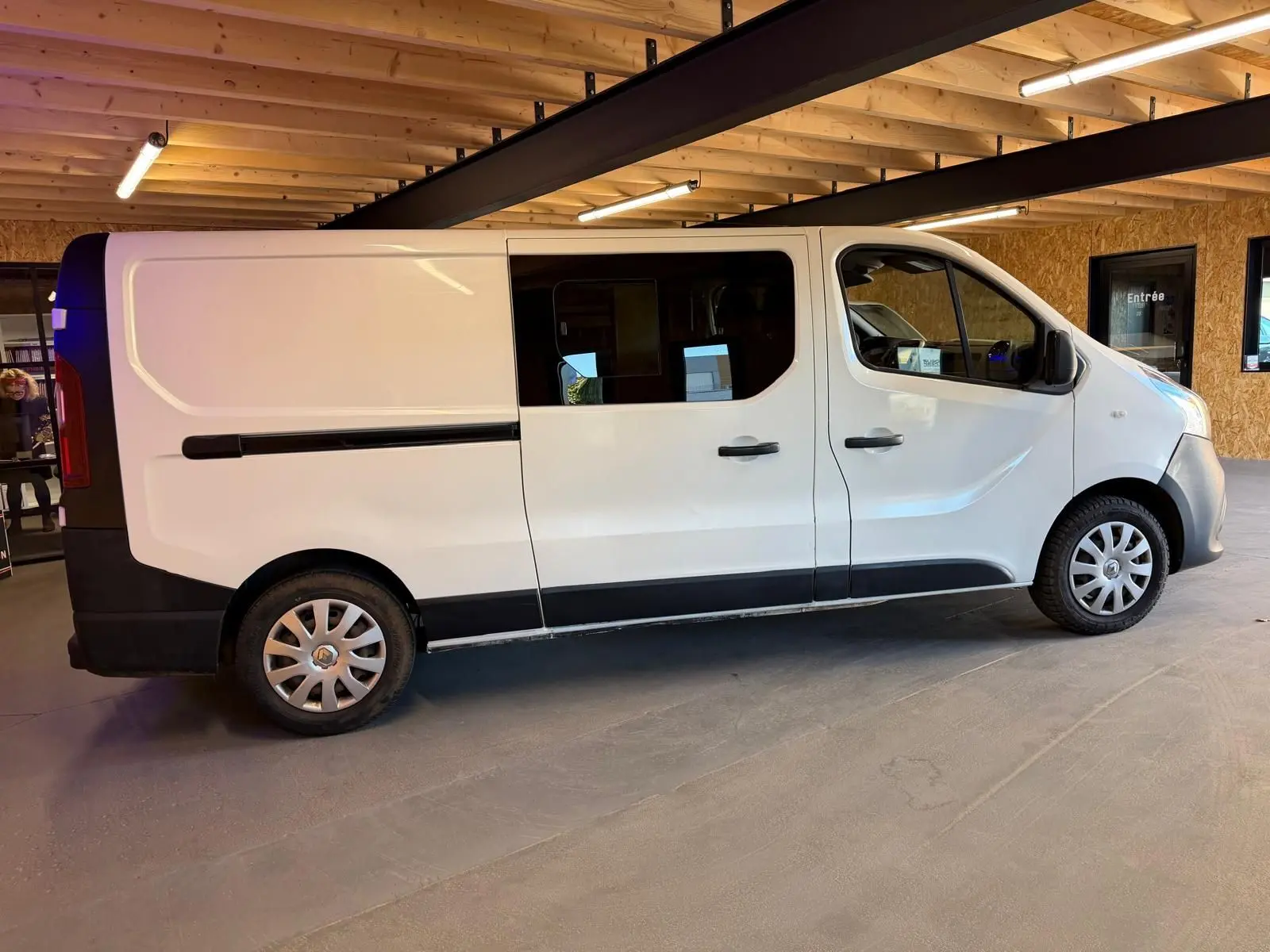 Vue de profil côté droit d'un Renault Trafic Fourgon blanc avec fenêtres latérales et jantes argentées, stationné en intérieur.