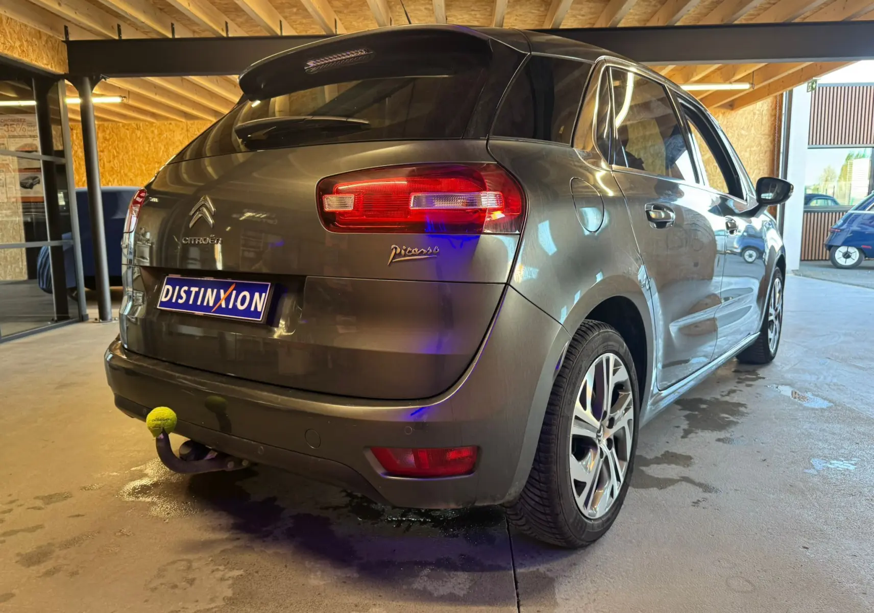 Vue 3/4 arrière droite d'une Citroën C4 Picasso gris métallisé avec attelage et jantes alliage en intérieur d'atelier.