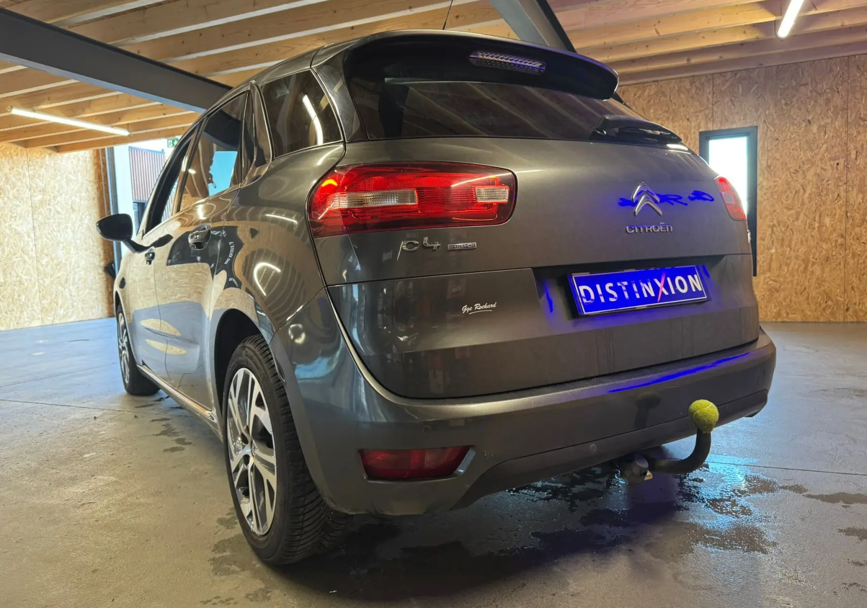 Vue 3/4 arrière gauche d'une Citroën C4 Picasso gris métallisé avec attelage et jantes alliage en intérieur garage.