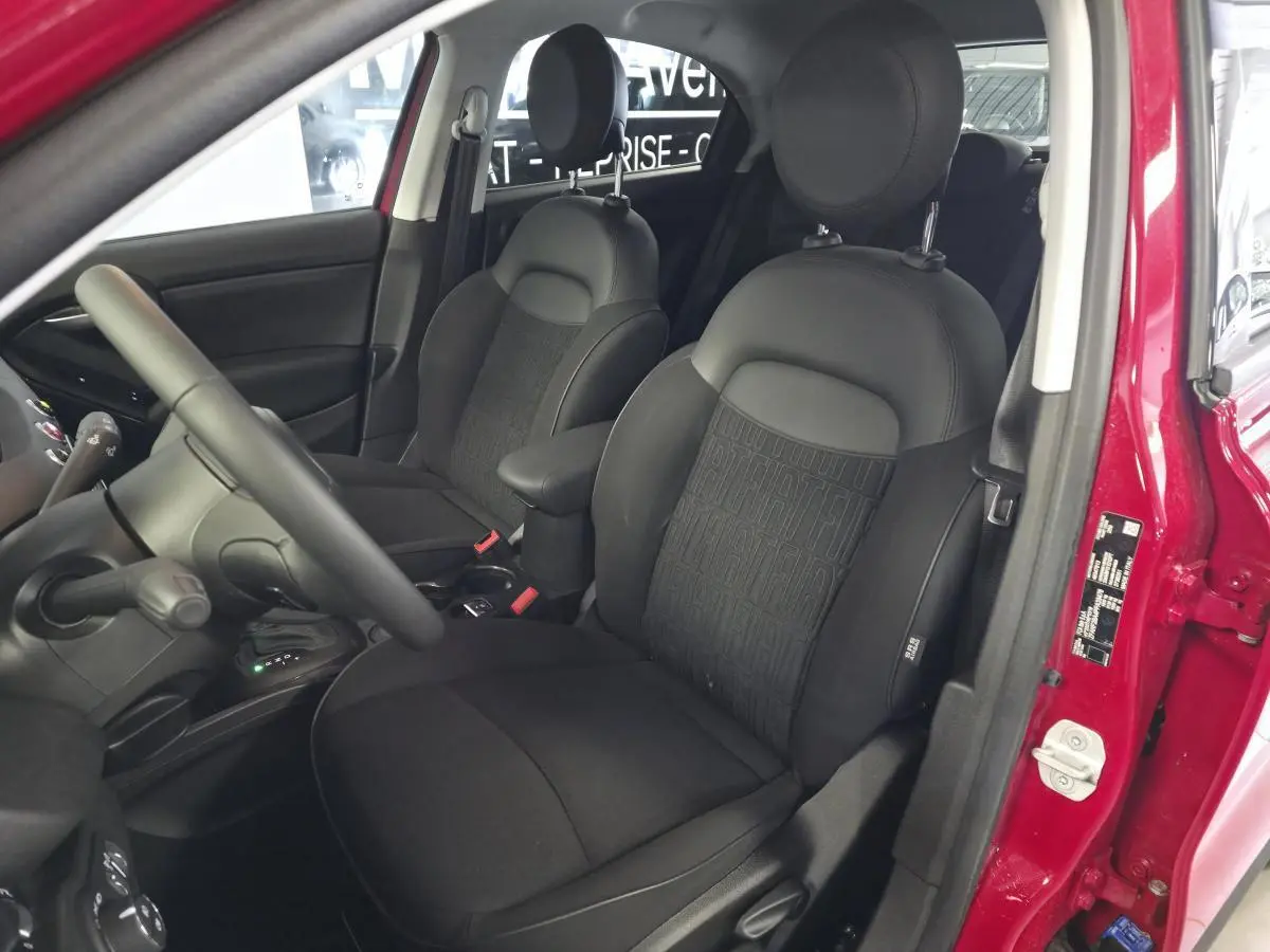 Vue intérieure côté conducteur de la Fiat 500X 2024 rouge, sièges tissu noir avec monogramme FIAT et accoudoir central.
