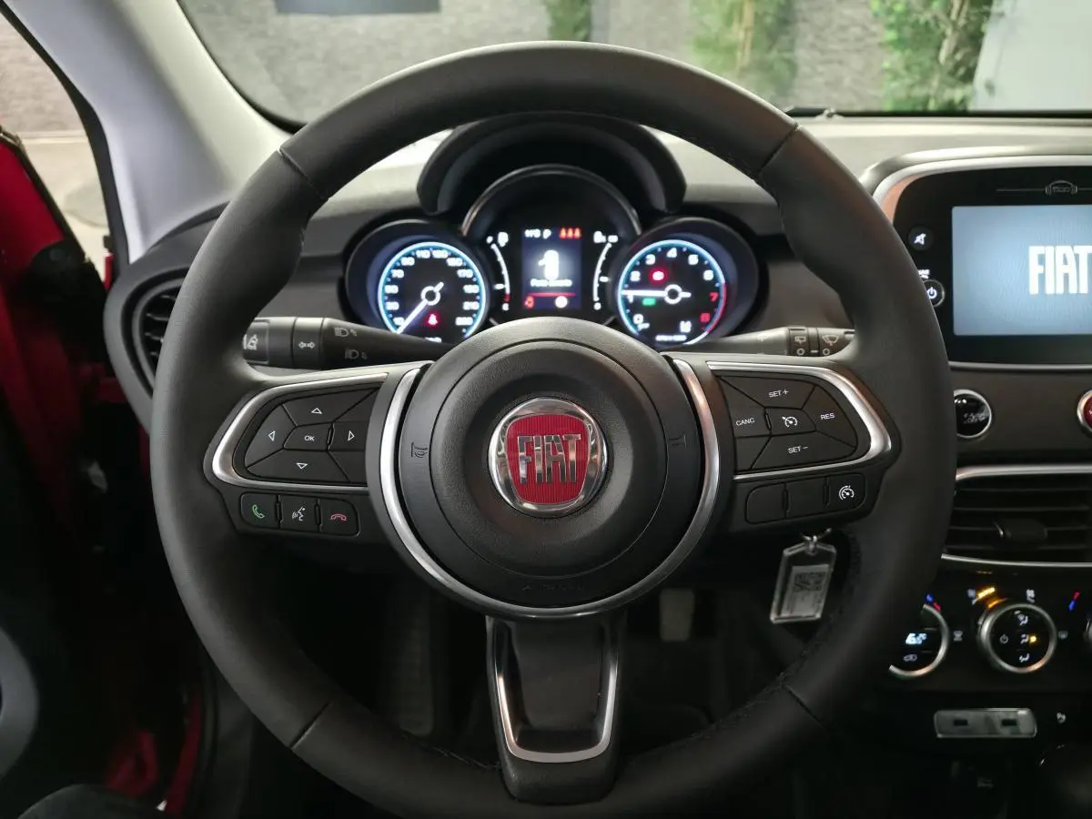 Vue intérieure centrée sur le volant noir soft-touch avec logo FIAT rouge et tableau de bord éclairé du 500X rouge.