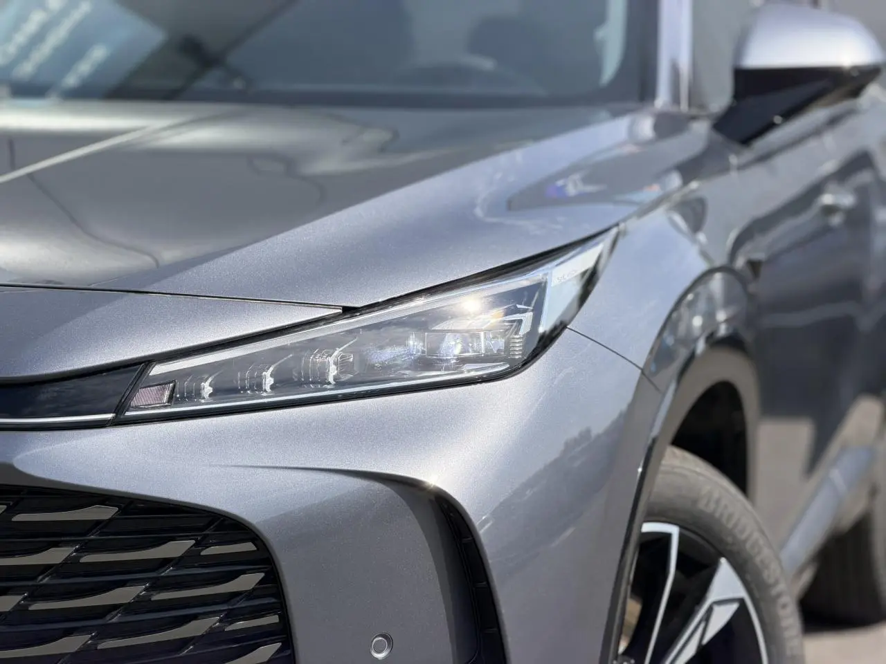 Gros plan sur l'avant droit gris métallisé du MG MOTOR EHS 1.5 Hybrid+ 224 Luxury avec phare LED et jante alliage 19 pouces.