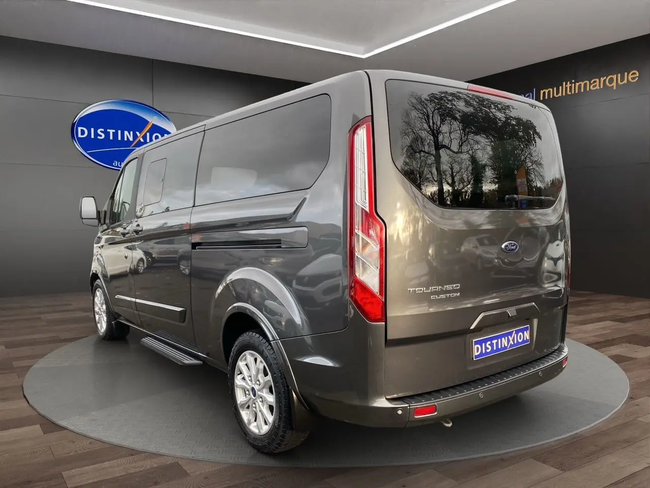 Vue 3/4 arrière droite d'un Ford Tourneo Custom gris magnetic avec hayon et vitres teintées en showroom.
