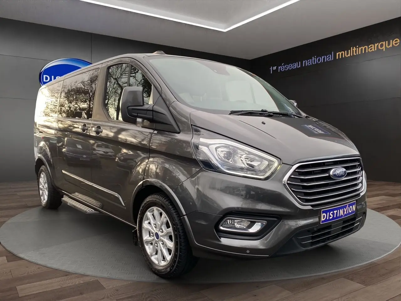 Ford Tourneo Custom gris Magnetic en 3/4 avant droit, avec calandre chromée et jantes alliage visibles.