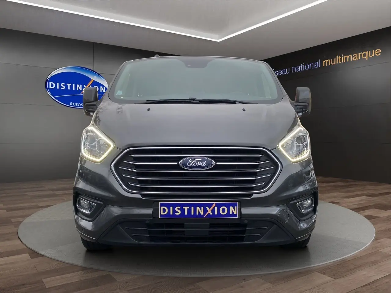 Vue frontale du Ford Tourneo Custom gris Magnetic avec phares allumés et logo Distinxion visible en arrière-plan.