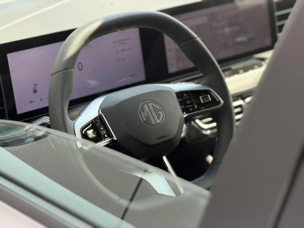 Gros plan sur le volant noir perforé avec logo MG, tableau de bord numérique et écran tactile en arrière-plan.