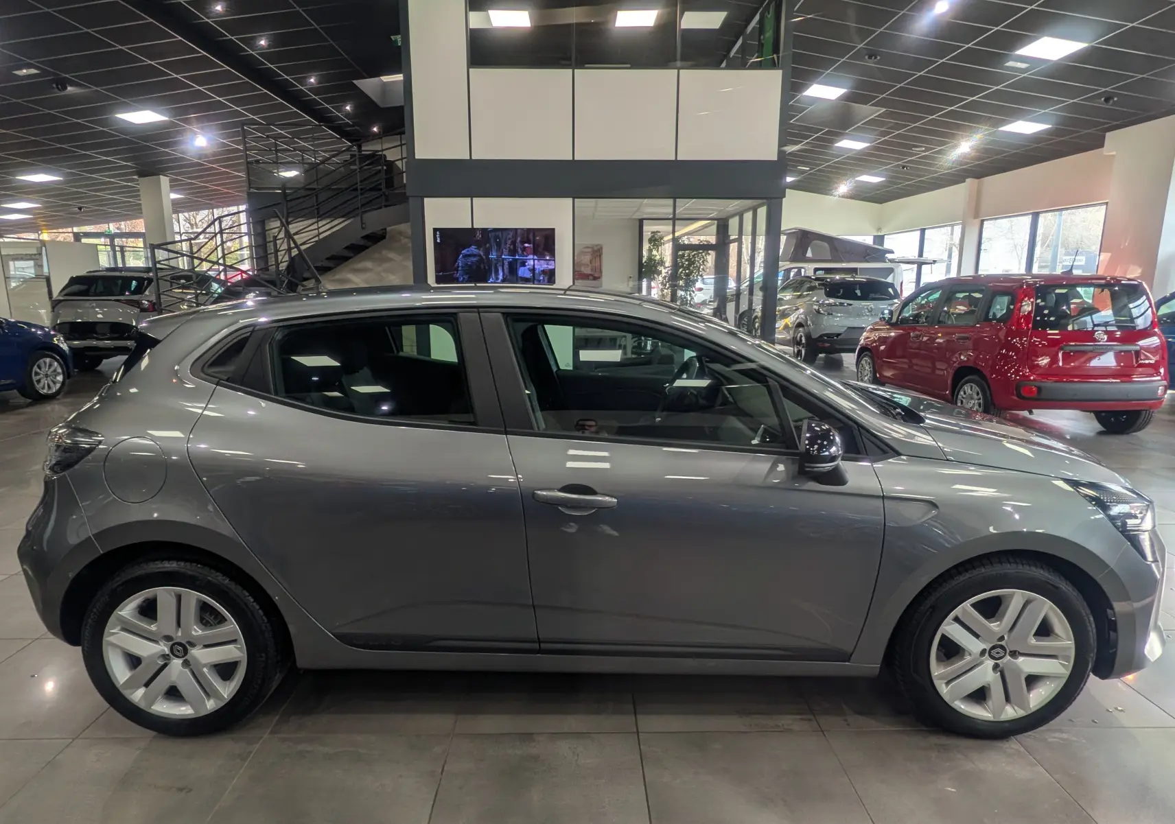 Profil côté gauche d'une Renault Clio V TCe 90 Evolution gris schiste, en intérieur de showroom avec jantes alliage.