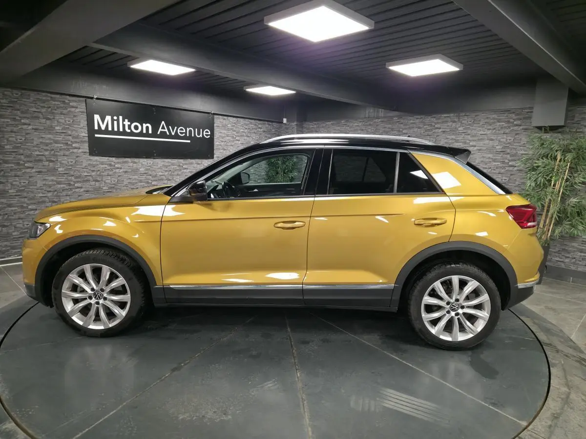 Vue latérale droite d'un Volkswagen T-Roc jaune Curcuma avec toit noir et jantes alliage 18 pouces dans un showroom.