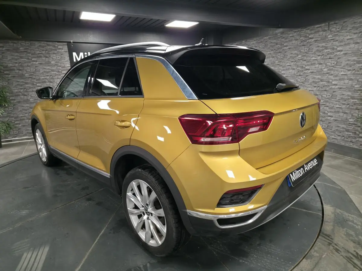 Vue 3/4 arrière droite d'un Volkswagen T-Roc jaune Curcuma avec toit noir et jantes alliage 18 pouces en intérieur showroom.