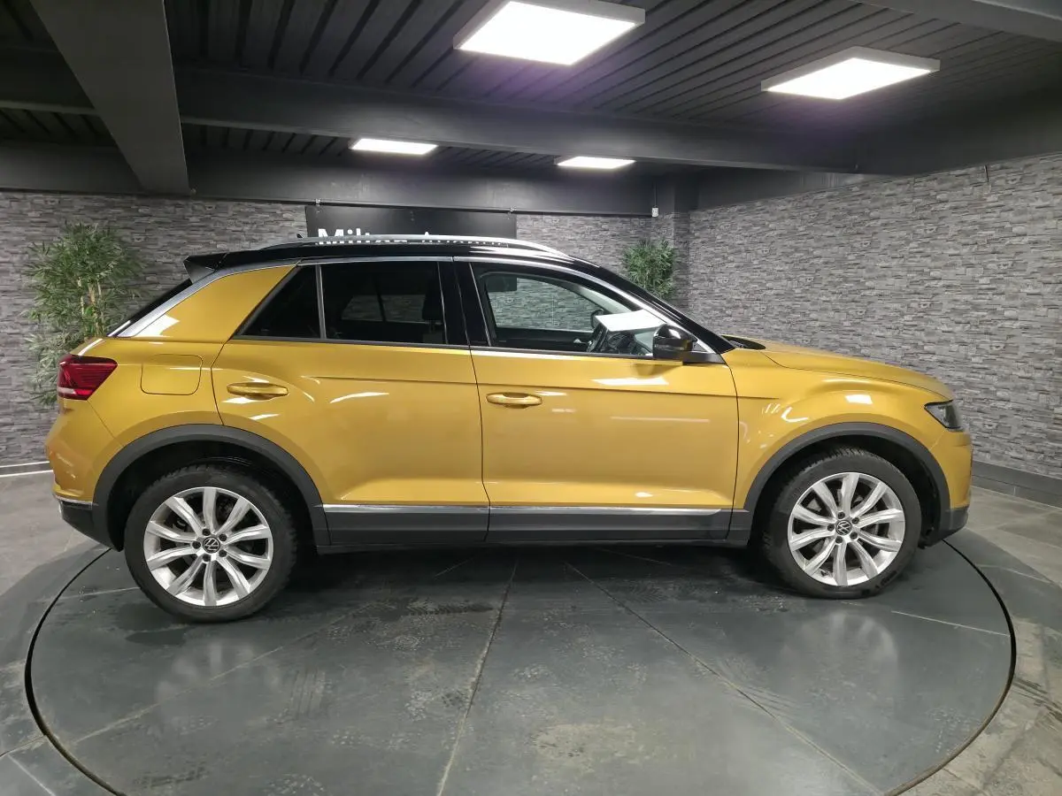 Vue de profil côté gauche d'un Volkswagen T-Roc jaune Curcuma avec toit noir et jantes alliage 18 pouces en intérieur showroom.